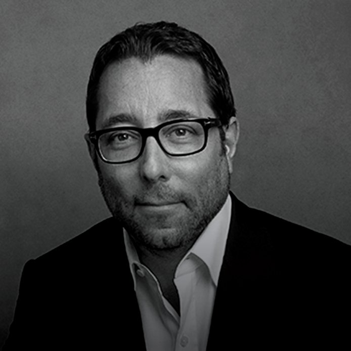 Black-and-white portrait of a man with glasses, wearing a suit and white shirt, looking at the camera with a slight smile.
