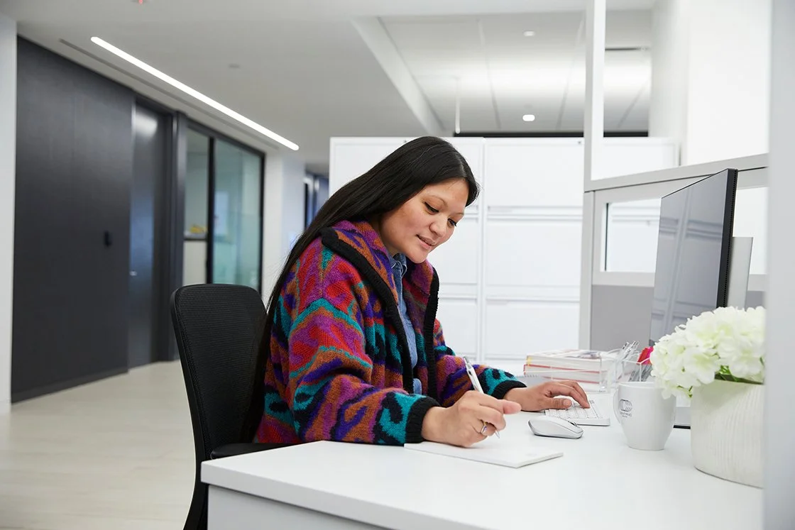 A woman sitting at a white desk in a modern office, writing on a notepad with a computer monitor, keyboard, mouse, and a white flower in a pot on the desk.