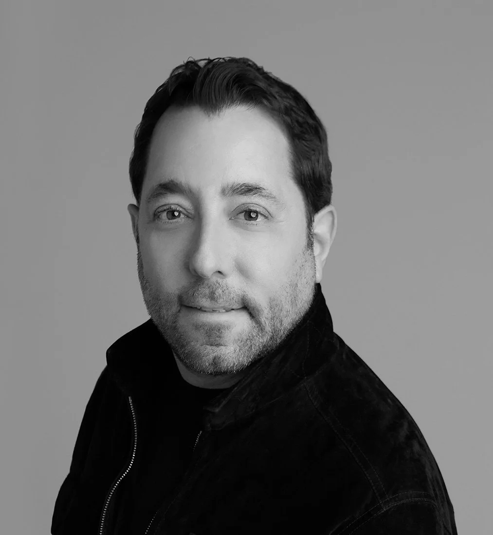 Black-and-white portrait of a man with glasses, wearing a suit and white shirt, looking at the camera with a slight smile.