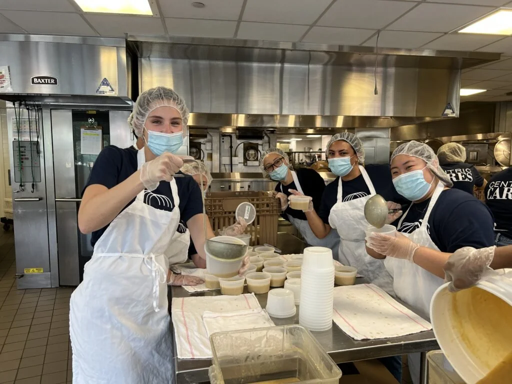 Group of people wearing masks, hairnets, and aprons working in a commercial kitchen, filling bowls with soup or similar food.