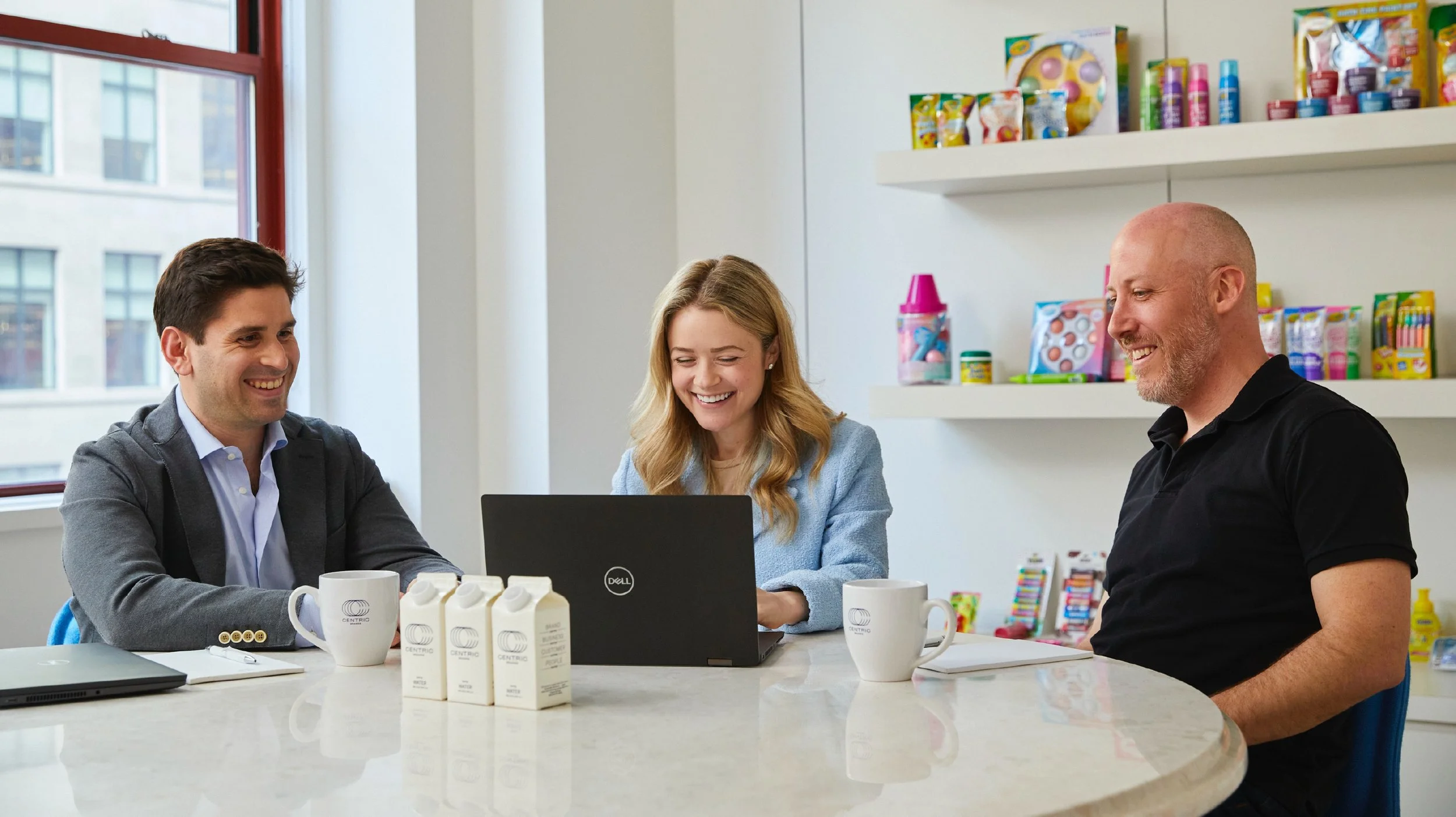Three people sitting at a table in an office or cafe, smiling and looking at a laptop, with drinks and milk cartons on the table, shelves with colorful items in the background.