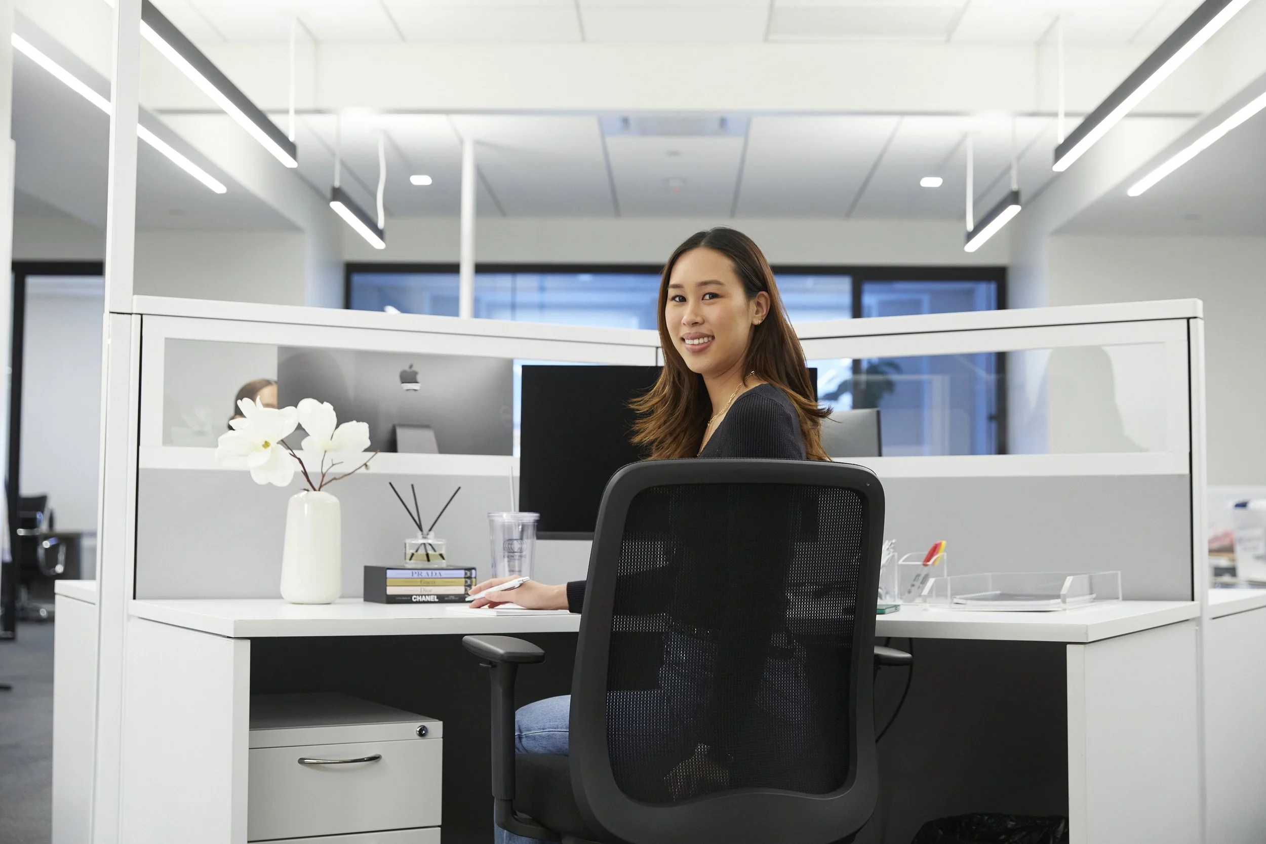 A woman sitting at her desk in an office, smiling, with a glass of water, decorative flower, and bookstore in front of her. She is seated in front of an Apple monitor and there are partition walls around her workspace.