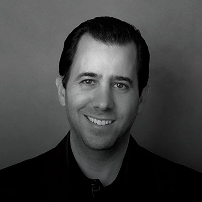 Black and white portrait of a smiling man with slicked-back hair wearing a dark collared shirt.