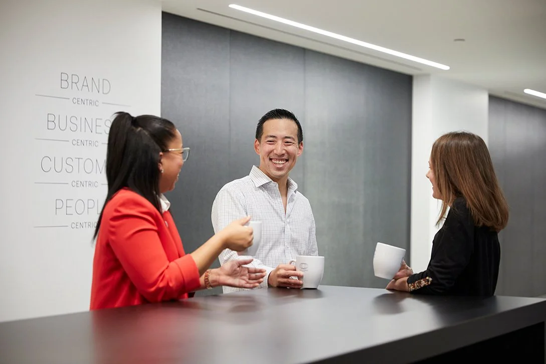 Three people smiling and talking with coffee mugs in an office lobby.
