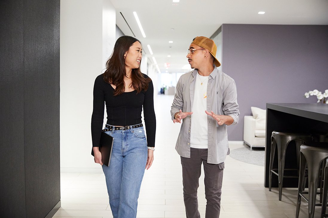 Two people, a woman and a man, are walking and talking indoors in a modern, brightly lit space. The woman is holding a tablet and smiling, while the man is gesturing with his hands.