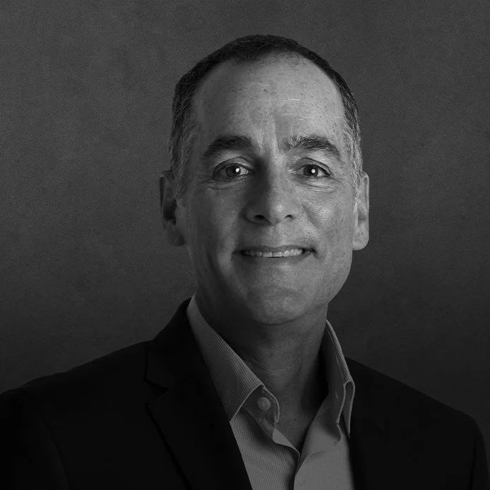 Black and white photo of a middle-aged man wearing a suit jacket and collared shirt, smiling slightly, with a plain dark background.