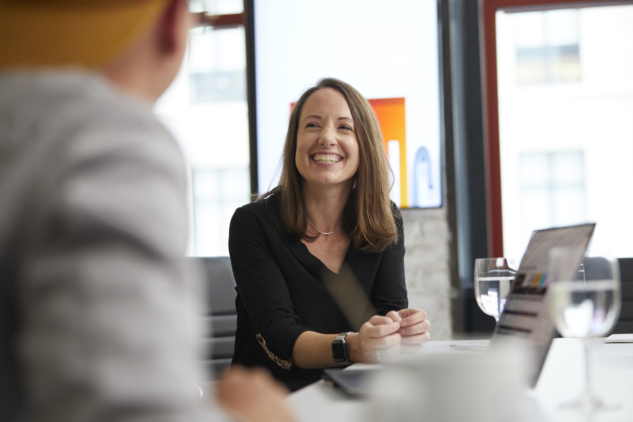 A woman sitting at a conference table smiling and talking to someone off-camera in a modern office environment with large windows, a laptop, glasses of water, and a large screen in the background.