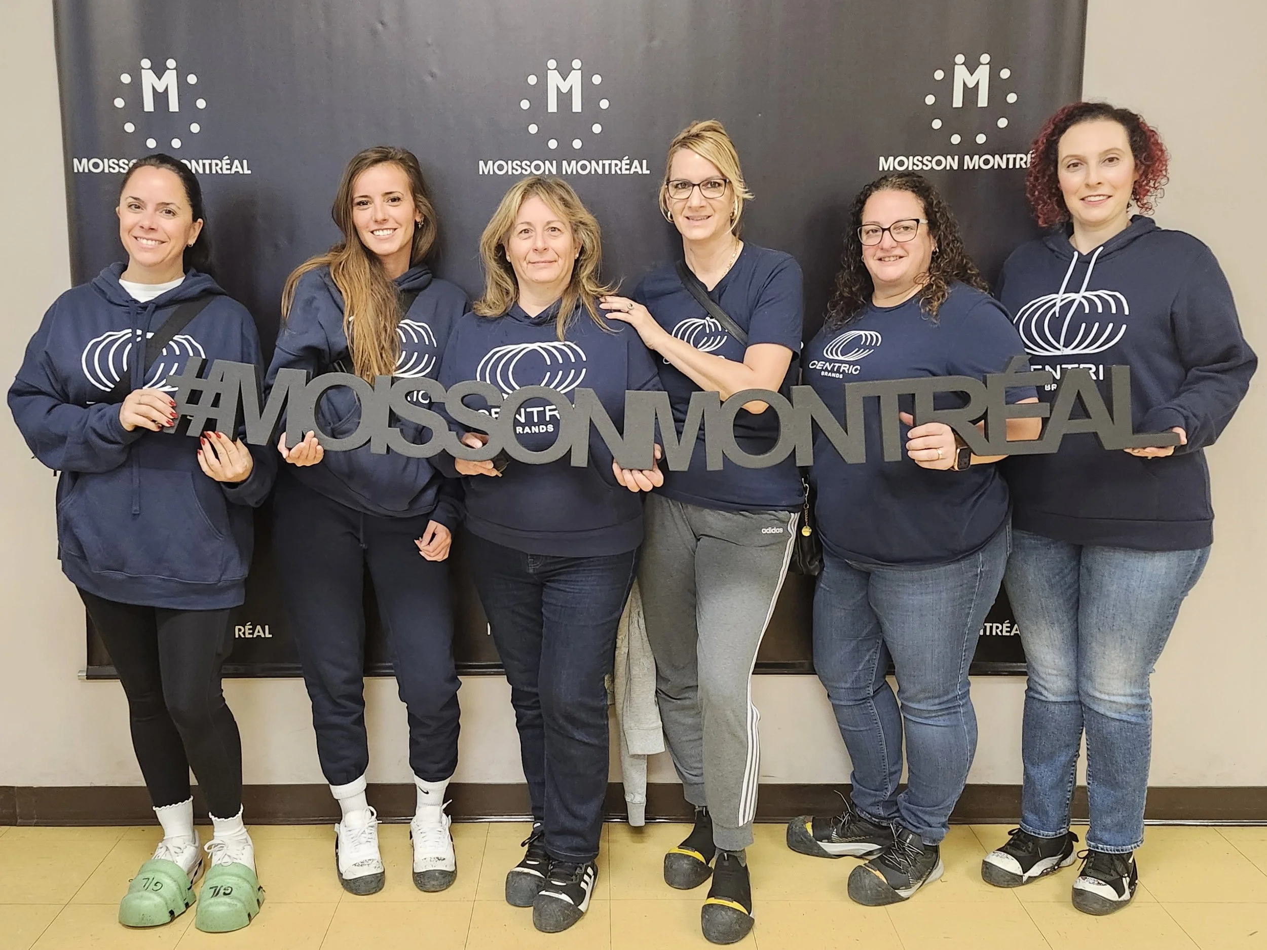 Group of six women wearing matching blue hoodies with a rainbow logo, holding a sign that says '#MOSSOMONTREAL,' standing in front of a black backdrop with 'MOISSON MONTRÉAL' written on it.