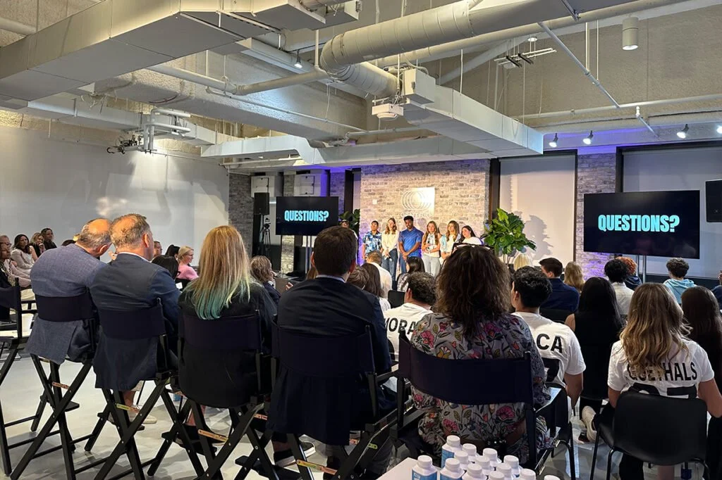 Audience attending a presentation or panel discussion in a modern, industrial-style venue with exposed pipes and large screens displaying the word 'QUESTIONS?'. The audience includes diverse adults and students, some wearing school-themed shirts.
