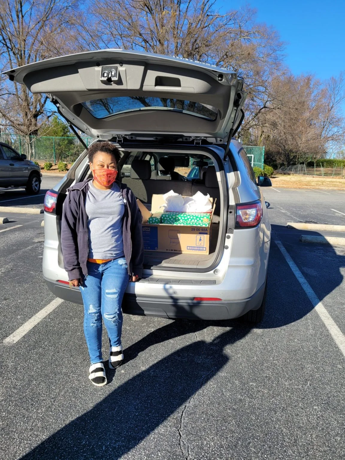 A woman stands next to a silver SUV with the trunk open, filled with boxes, in a parking lot on a sunny day. She is wearing a red face mask, a gray t-shirt, a black jacket, ripped jeans, and black and white sandals.