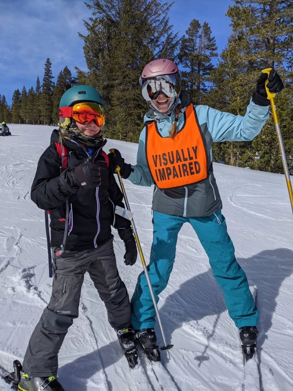 Two women on skis standing in the snow with a forested mountain background. One woman wears a black ski jacket and helmet with orange goggles, and the other wears a blue jacket and pink helmet with reflective goggles. The woman in blue has an orange vest that reads 'Visually Impaired' and is holding a yellow ski pole.