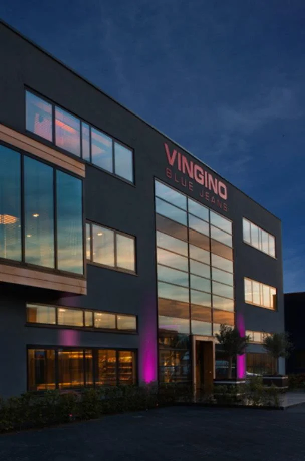 Exterior of VINGINO Blue Jeans store during sunset, with purple lighting illuminating the lower part of the building and large glass windows reflecting the sky.