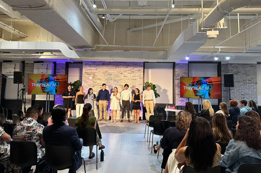A group of eight people on stage in front of an audience, with two large screens displaying colorful images and text that says 'They're All AI' at an indoor conference or event space.