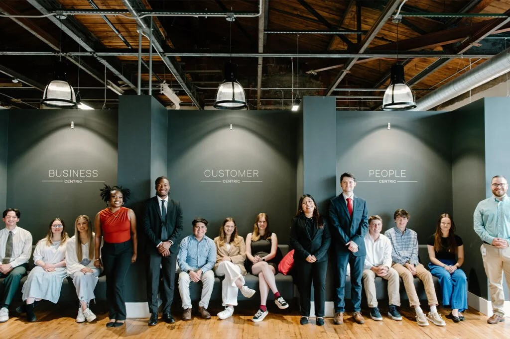 A diverse group of young adults dressed in business casual attire, standing and sitting in a modern office space with dark green walls labeled 'BUSINESS CENTRIC,' 'CUSTOMER CENTRIC,' and 'PEOPLE CENTRIC.'