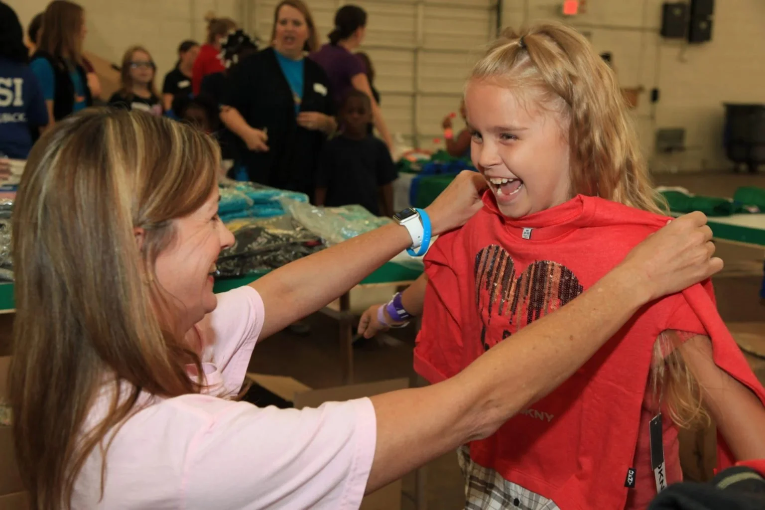 A woman and a girl are sharing a joyful moment at an indoor event. The woman, smiling with eyes closed, is placing a red shirt over the girl's shoulders. The girl has long blonde hair, is wearing a red shirt with a heart on it, and is laughing happily.