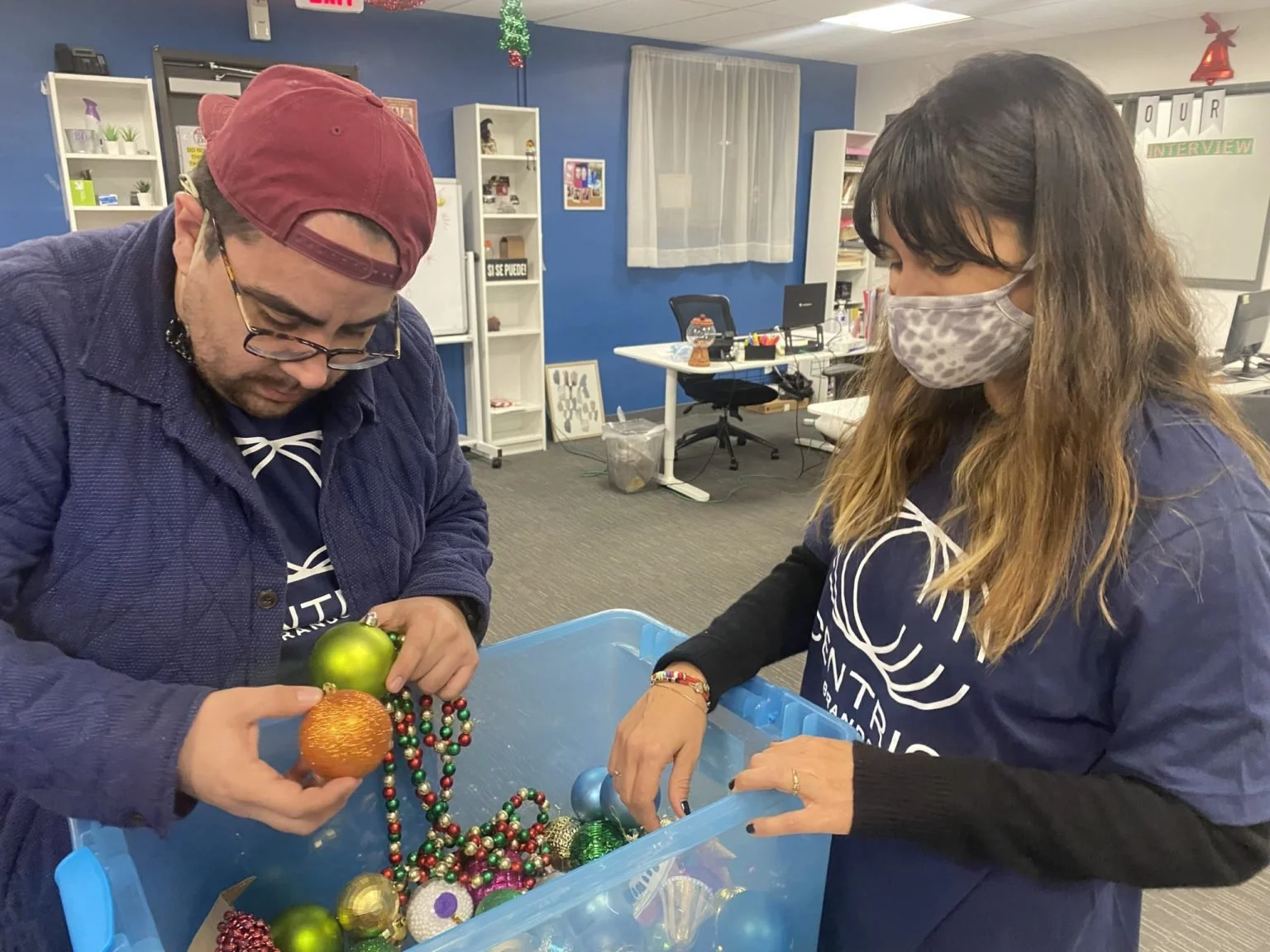 Two people decorating Christmas ornaments with a box of various colorful decorations in an office setting.