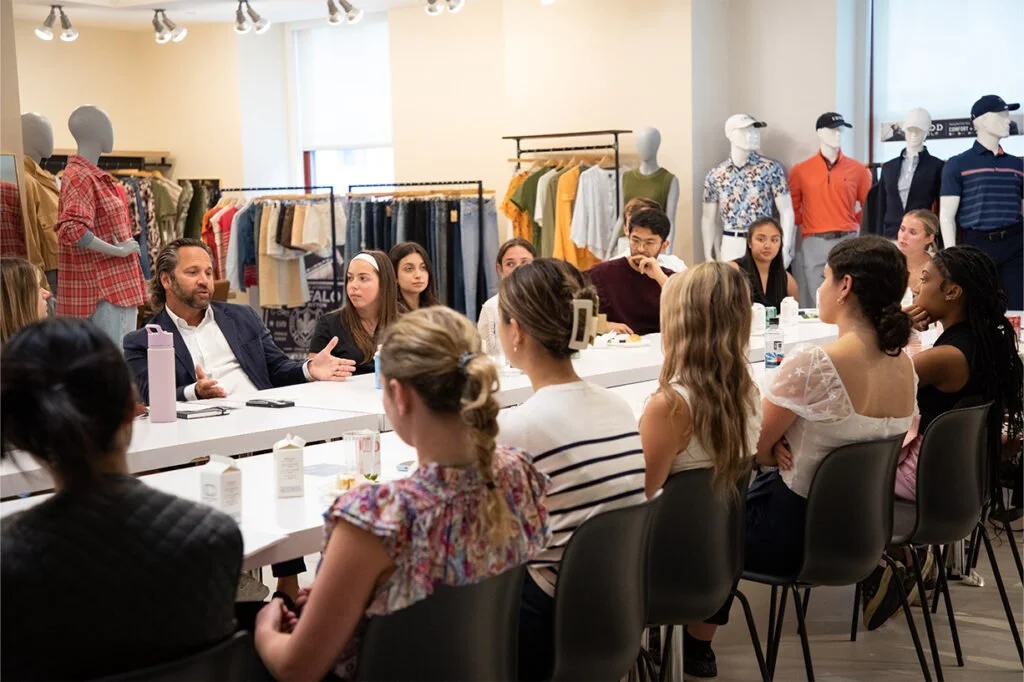 A group of people participating in a meeting or discussion in a clothing store or showroom, with mannequins and clothing displays in the background.