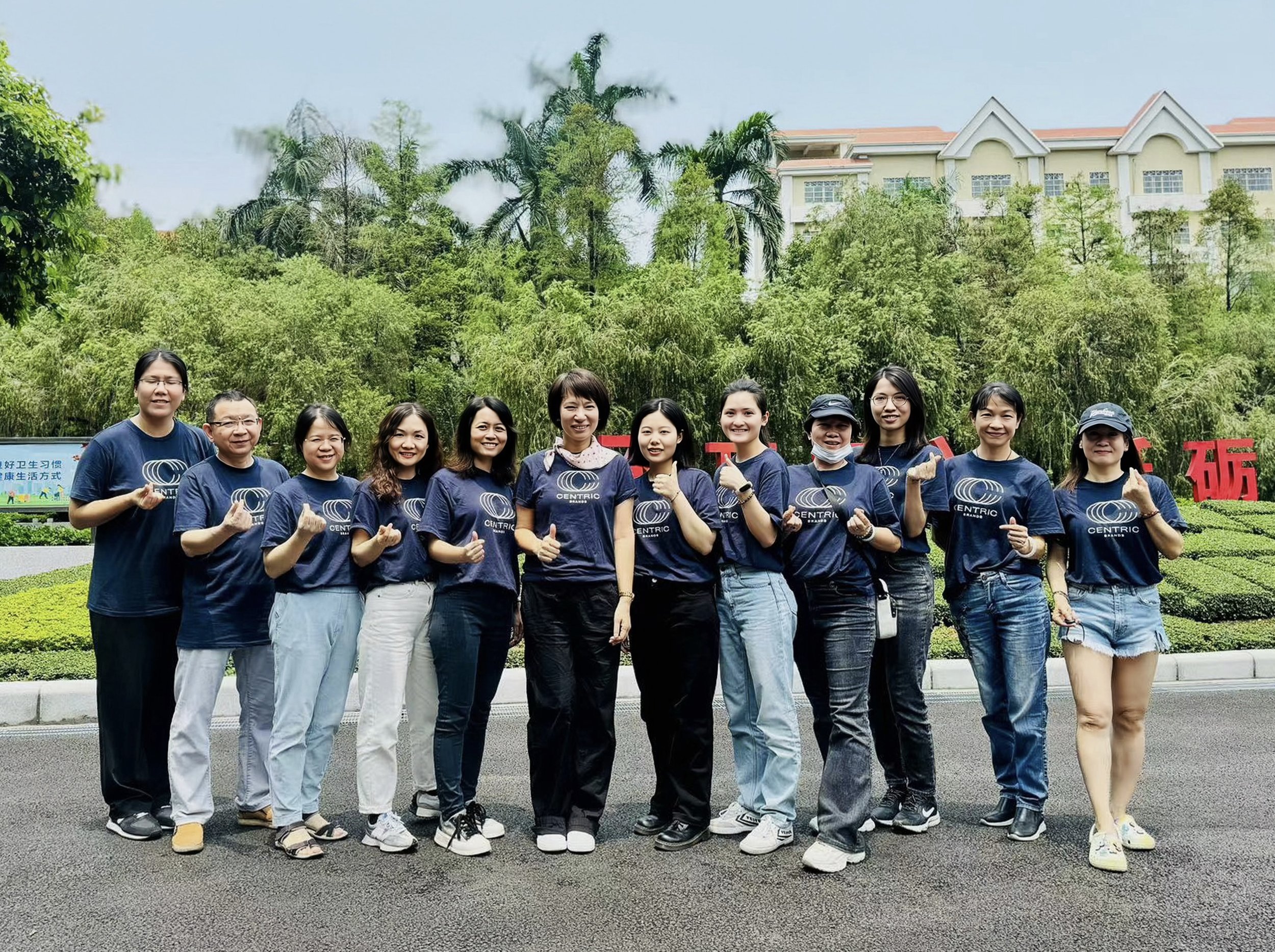 Group of eleven women standing on pavement, smiling and making peace signs, wearing matching navy blue T-shirts with a logo and the word 'CENTRIC' printed on them, in front of lush green trees and a building with white walls and red roof.