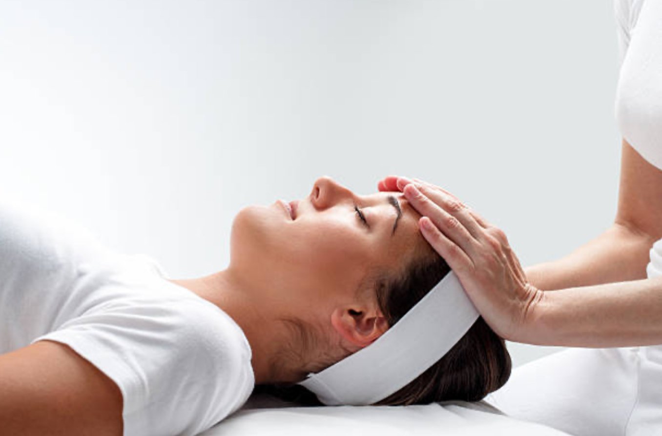 A woman is lying down comfortably with her eyes closed in a spa setting. The reiki practitioners hands are resting on her head. They are both dressed in white.