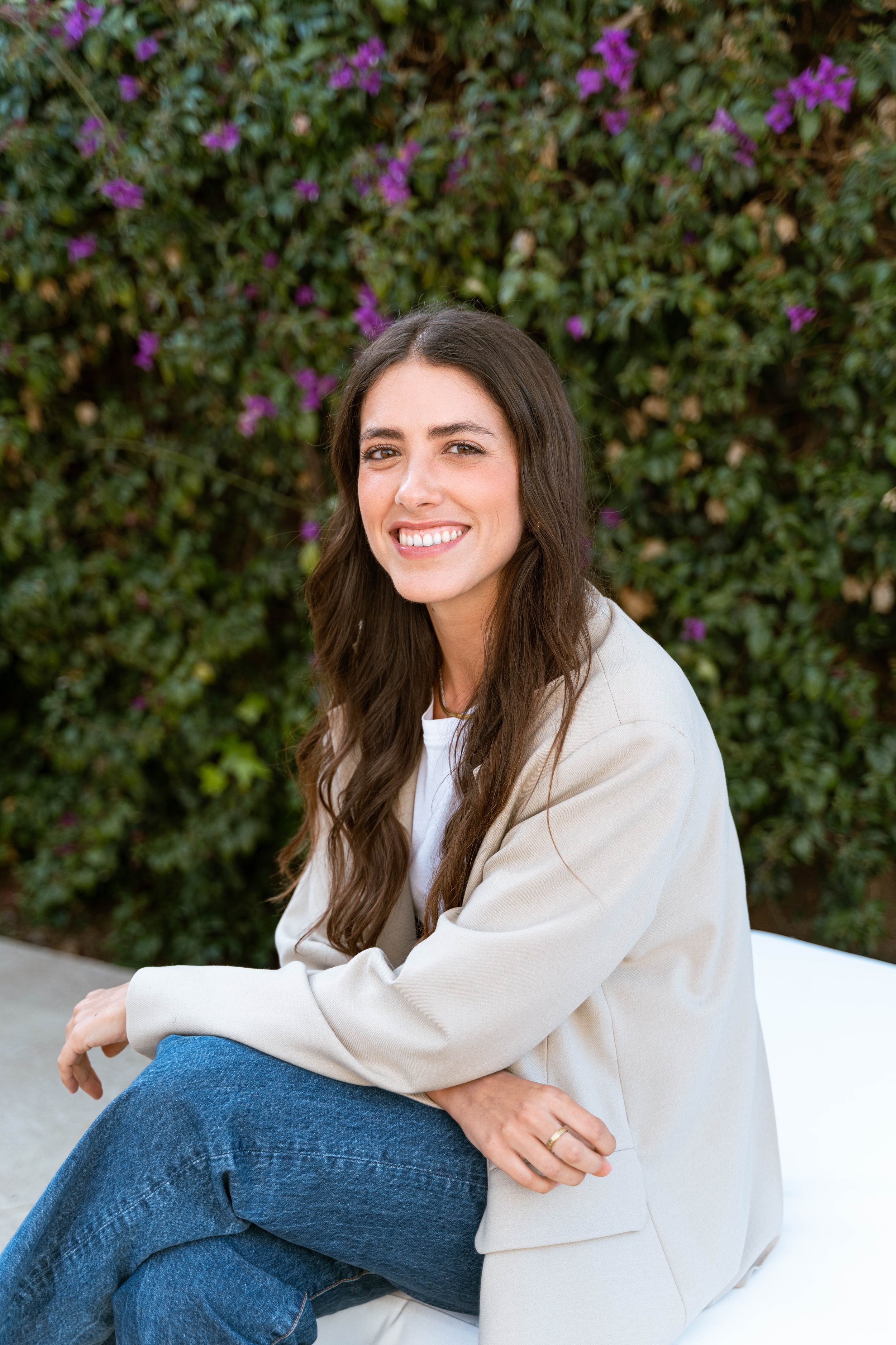 Smiling woman with long brown hair, wearing a white shirt, beige jacket, and jeans, sitting outdoors with green foliage in the background.