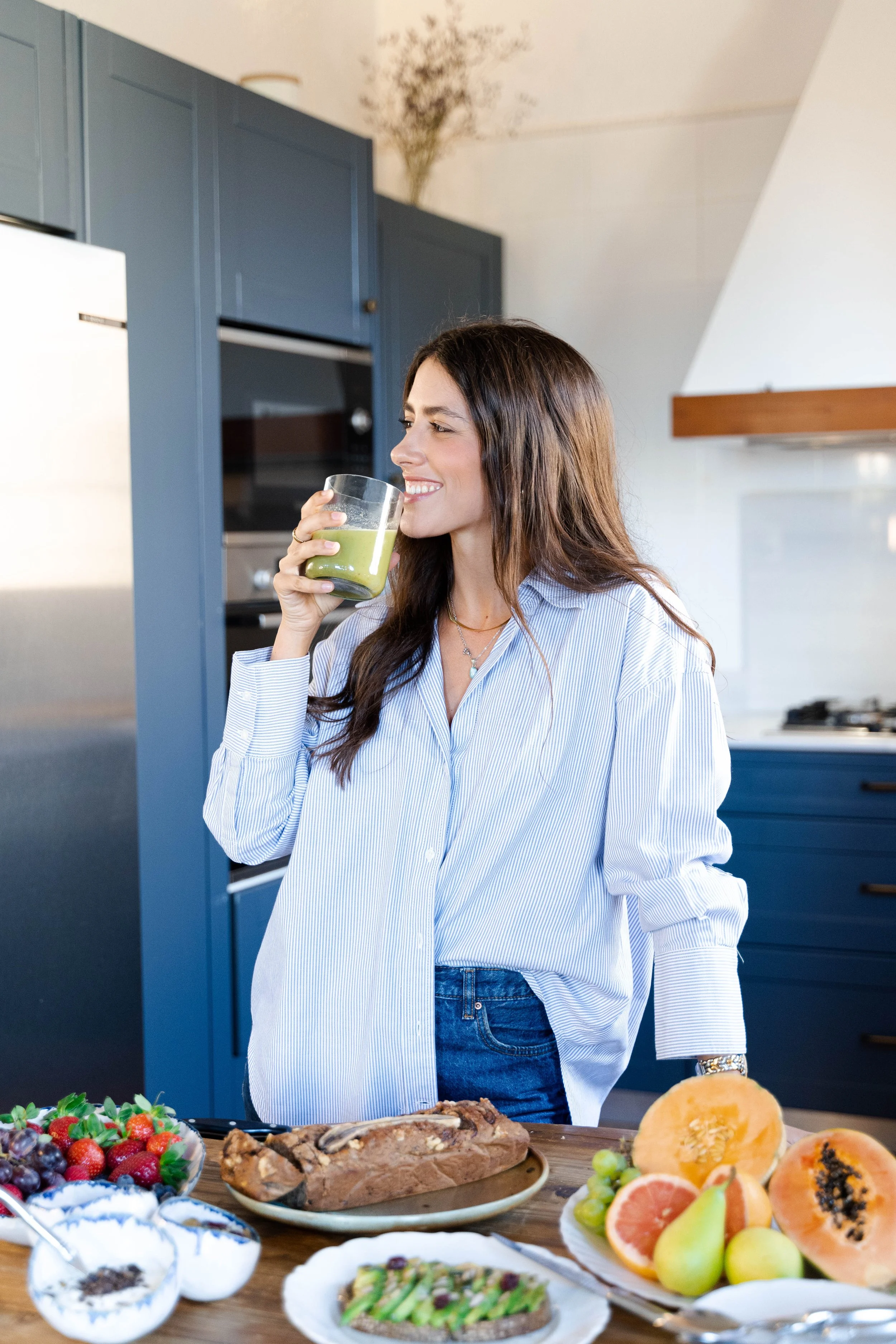 Woman in kitchen drinking green juice with plates of fruit and bread on table.