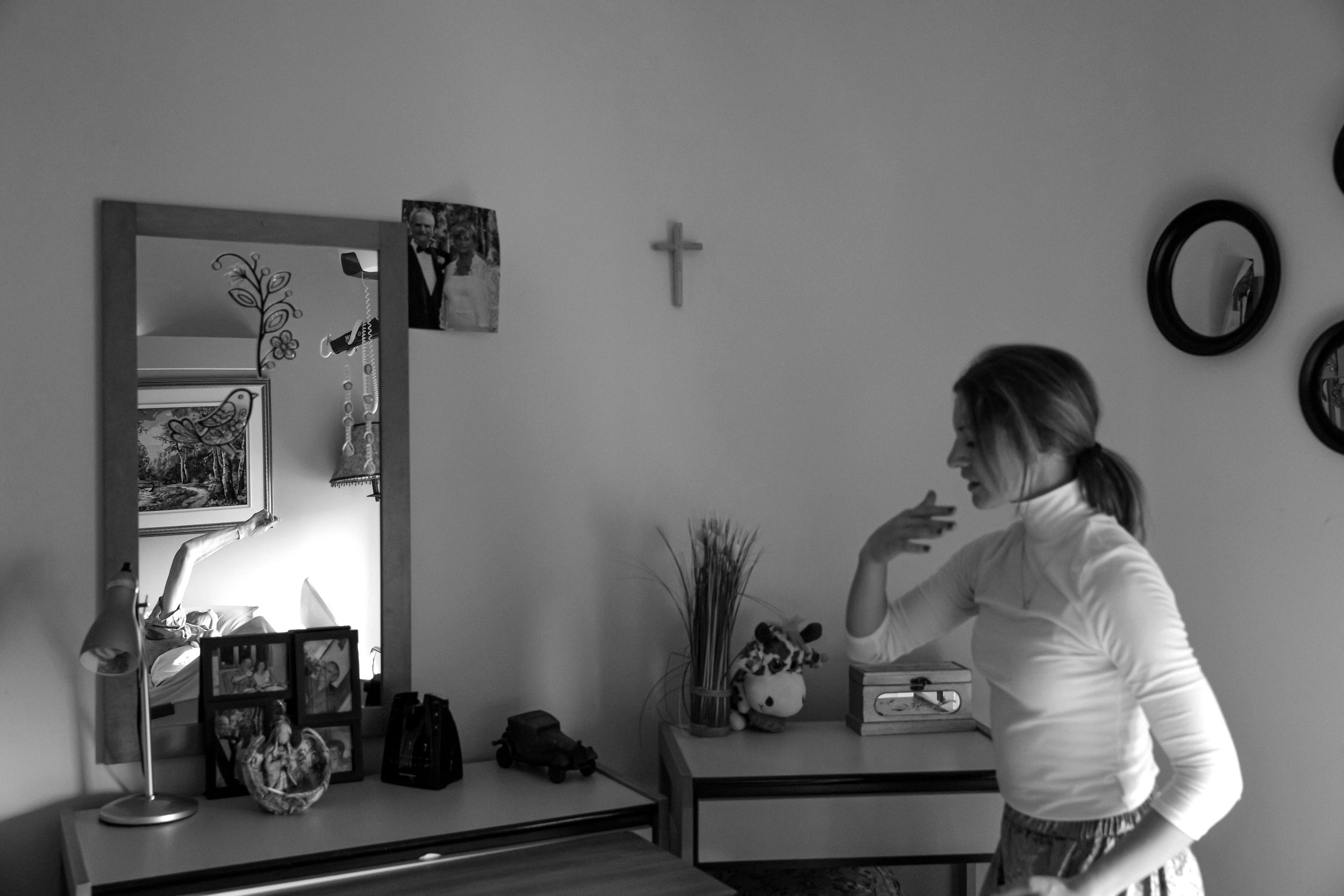 Une femme en robe blanche dans une pièce, regardant dans un miroir, avec des photos de famille et des décorations sur la table à côté, en noir et blanc.