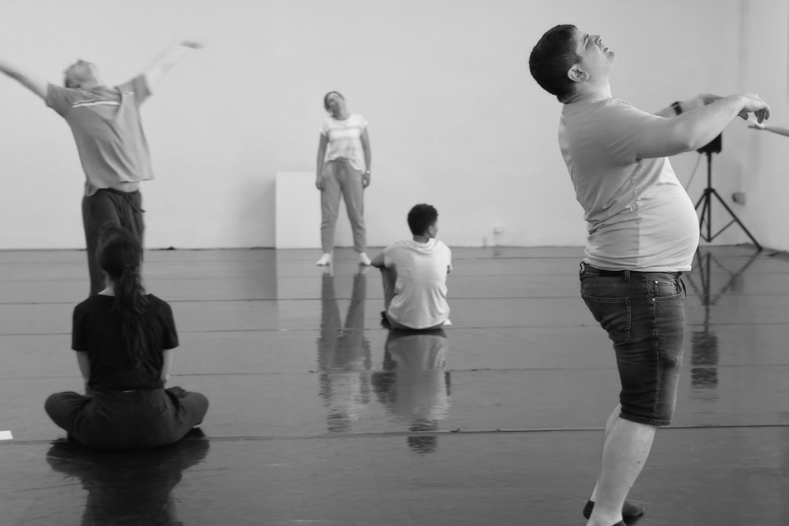 Groupe de personnes dans un studio de danse, certaines assises sur le sol, d'autres debout, en arrière-plan, en train de pratiquer ou d'observer, en noir et blanc.