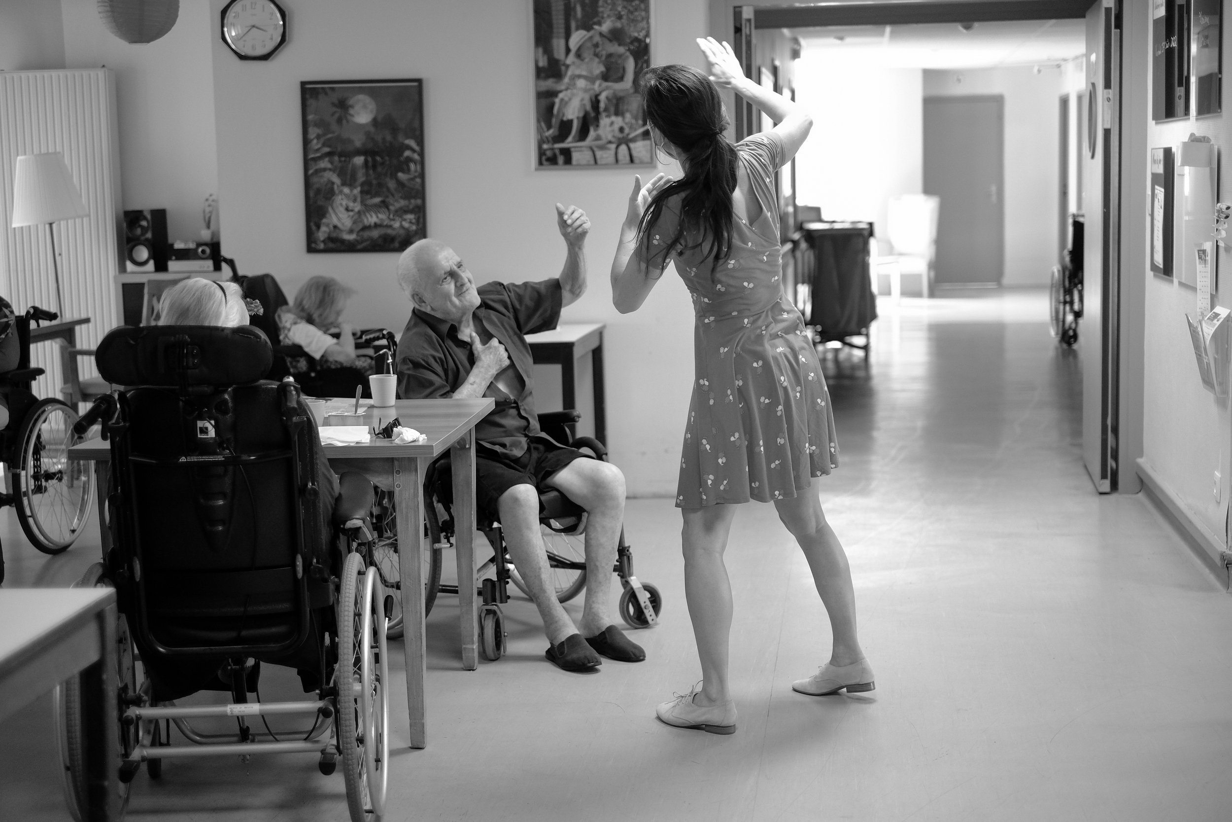 Une femme danse dans un couloir d'une maison de retraite, entourée de personnes âgées dans des fauteuils roulants, en noir et blanc.