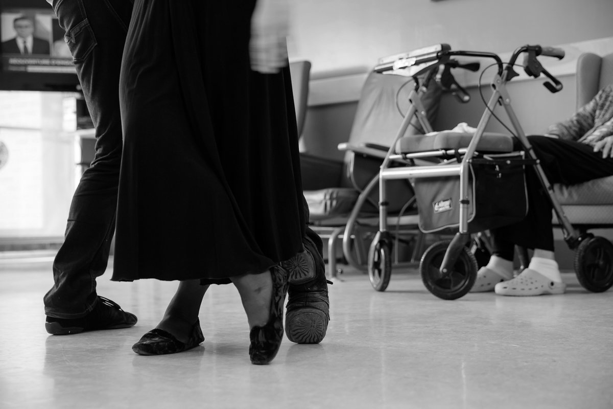 Deux personnes dans un centre de soins ou une école, une dansant avec des chaussures de danse, l'autre assise sur une chaise roulante.