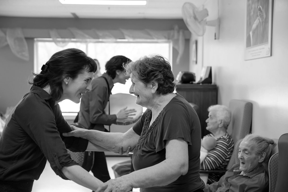 Une jeune femme et une vieille dame se sourient et se tiennent la main dans une pièce lumineuse, probablement dans une maison de retraite, entourées d'autres personnes âgées.