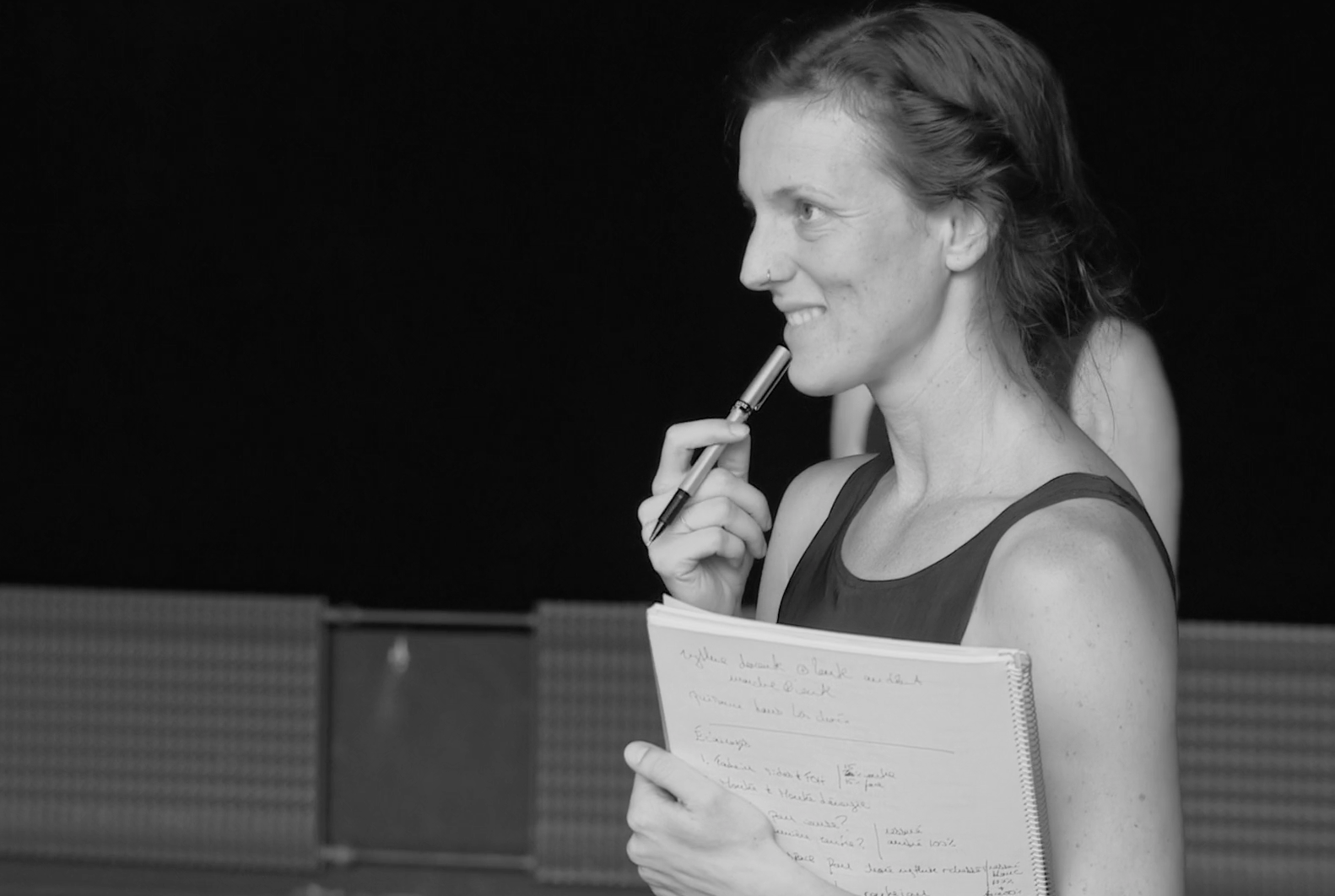 Une femme souriante, tenant un carnet avec des notes manuscrites, tenant un stylo près du visage, en tenue de sport, dans un cadre noir et blanc.