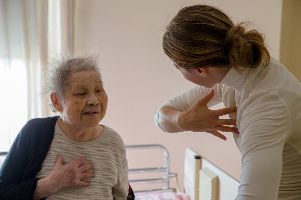 Une infirmière ou une personne en uniforme médical regarde une femme âgée qui a la main sur la poitrine, probablement en train de faire un exercice ou une évaluation médicale.