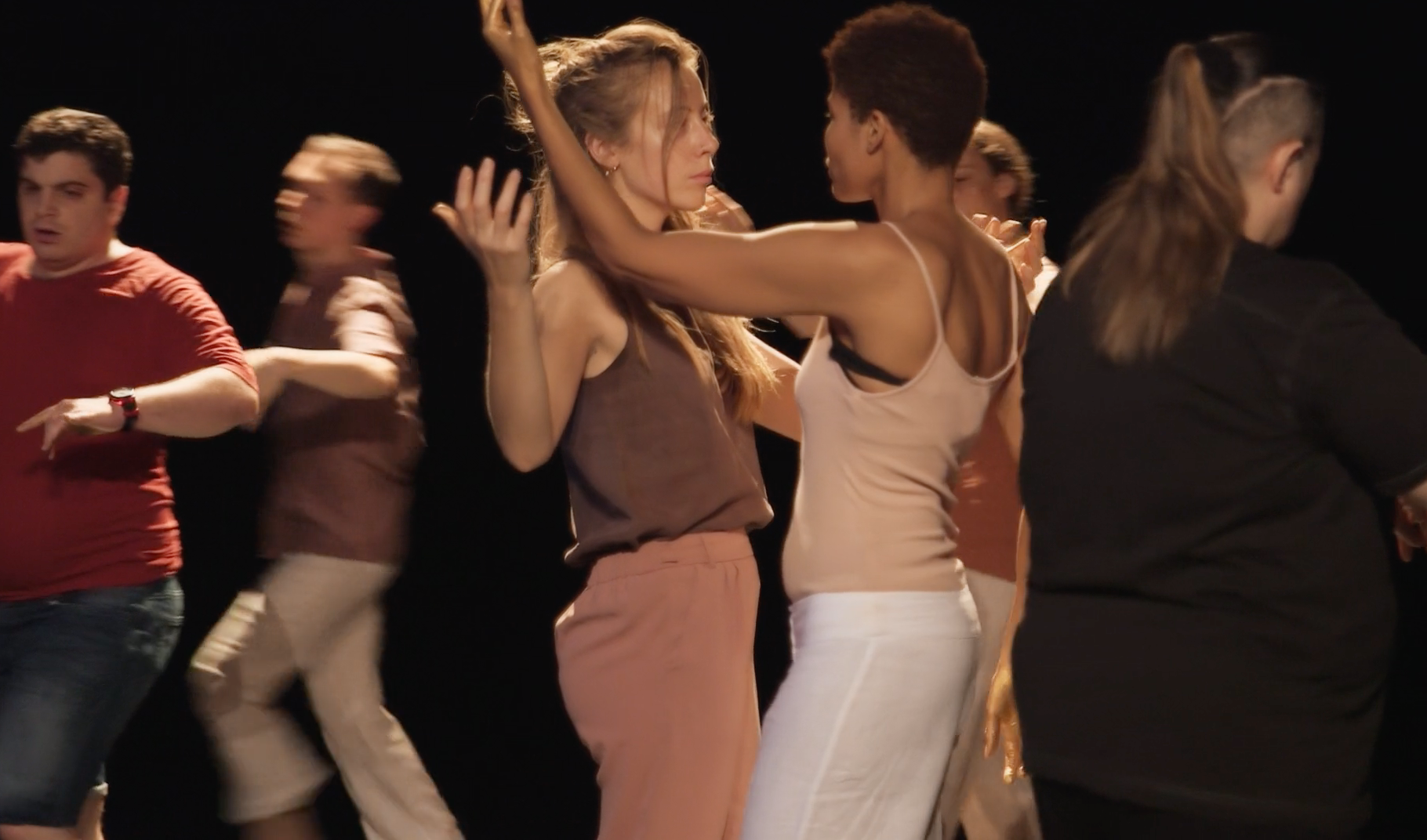Groupe de personnes dansant ensemble, hommes et femmes, dans un contexte de spectacle ou de répétition avec un fond noir.