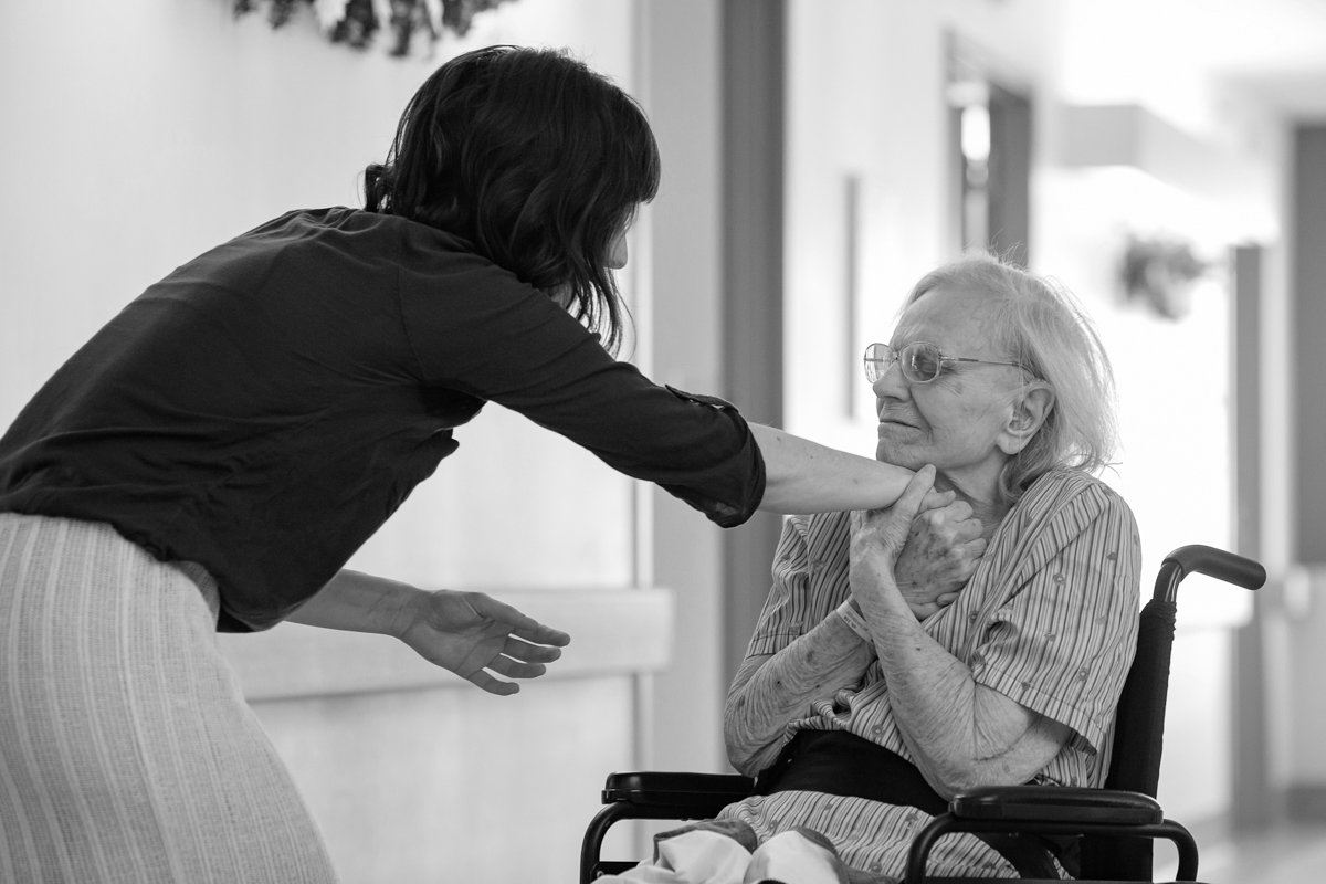 Une femme aidant une personne âgée en fauteuil roulant à se défendre ou se protéger, dans un cadre intérieur.