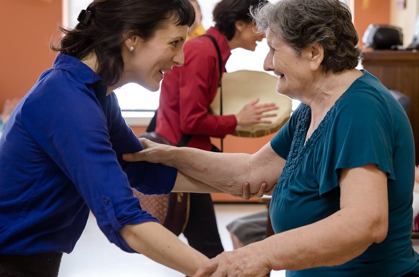 Une femme souriante aide une personne âgée dans un centre de soins ou une maison de retraite, elles se tiennent la main et échangent un regard chaleureux.