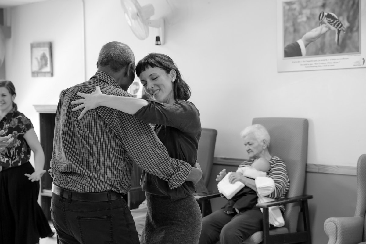 Deux personnes dansent dans une pièce, une femme souriante et un homme vu de dos, avec une femme âgée assise, tenant un bébé, en arrière-plan.