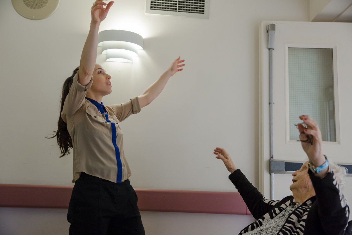 Une jeune femme aidant une personne âgée à faire des exercices de rééducation ou de thérapie en levant les bras dans une salle.