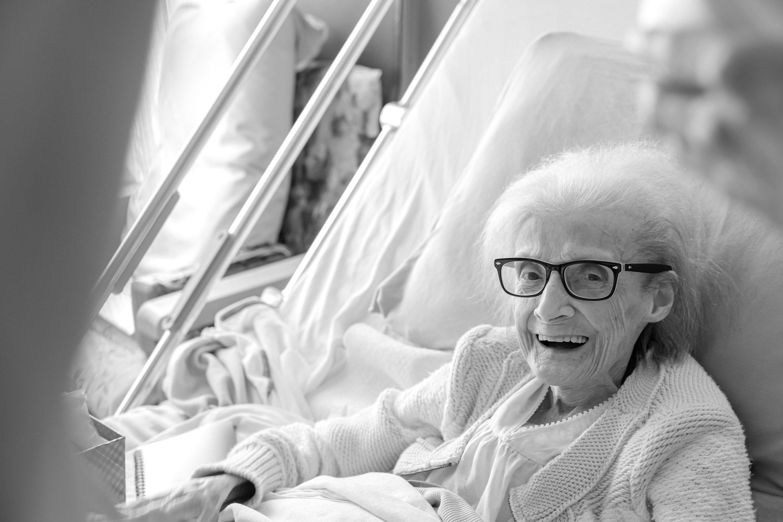 Une vieille femme souriante à l'hôpital, portant des lunettes et allongée dans un lit.