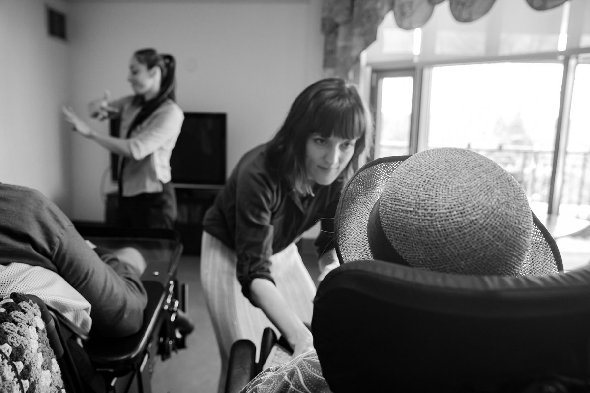 Une femme avec des cheveux bruns court, portant un haut noir et une jupe rayée, regarde vers l'appareil photo tandis qu'une autre femme assise, portant un chapeau en paille, est partiellement visible devant elle. En arrière-plan, une femme avec une q