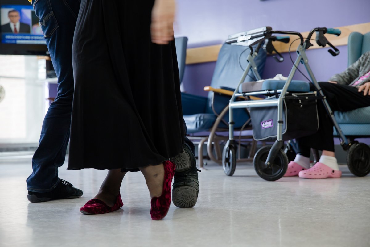 Une personne portant des chaussures rouges et une personne portant des chaussures noires sont dans un lieu semblant être un centre d'attente. À l'arrière, une personne assise dans un fauteuil roulant avec des chaussons roses. Des fauteuils et un déam