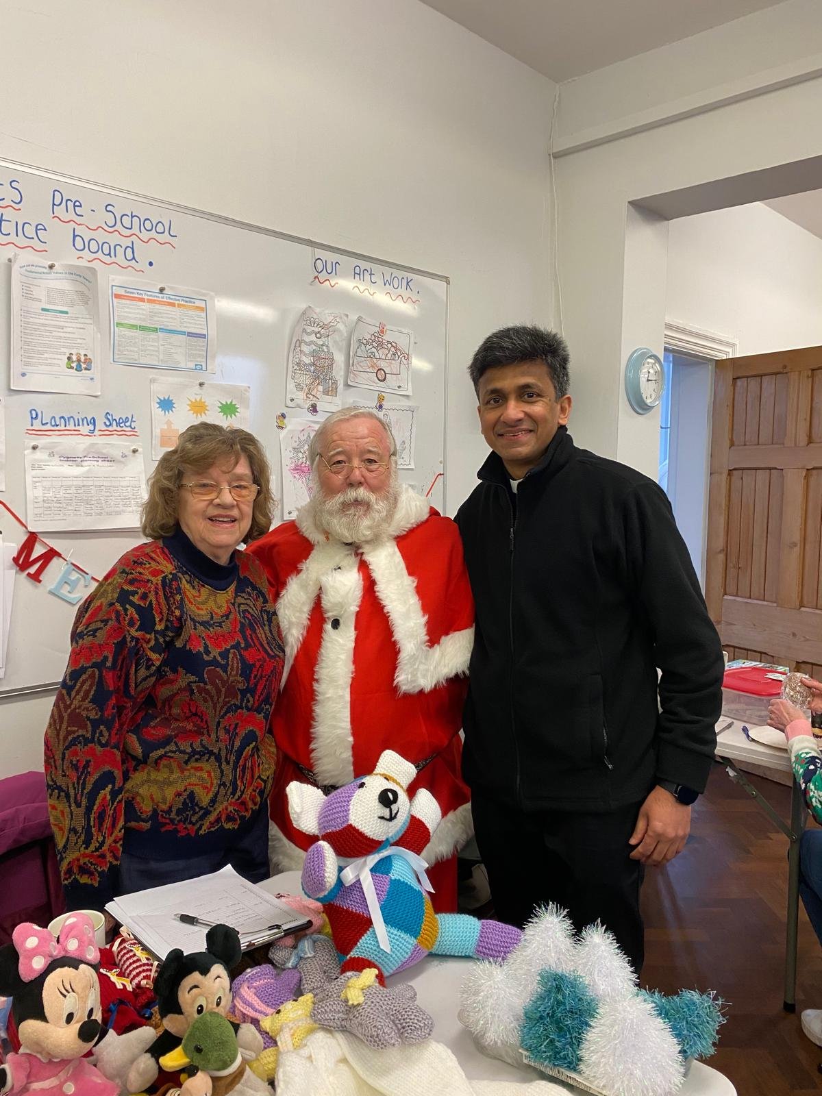 A group of clergy members dressed in red liturgical vestments standing during a religious ceremony inside a church. A young girl is visible in the back holding a book, and a colorful poster with a dove and words like 'LOVE,' 'PEACE,' 'KINDNESS' is in the background.