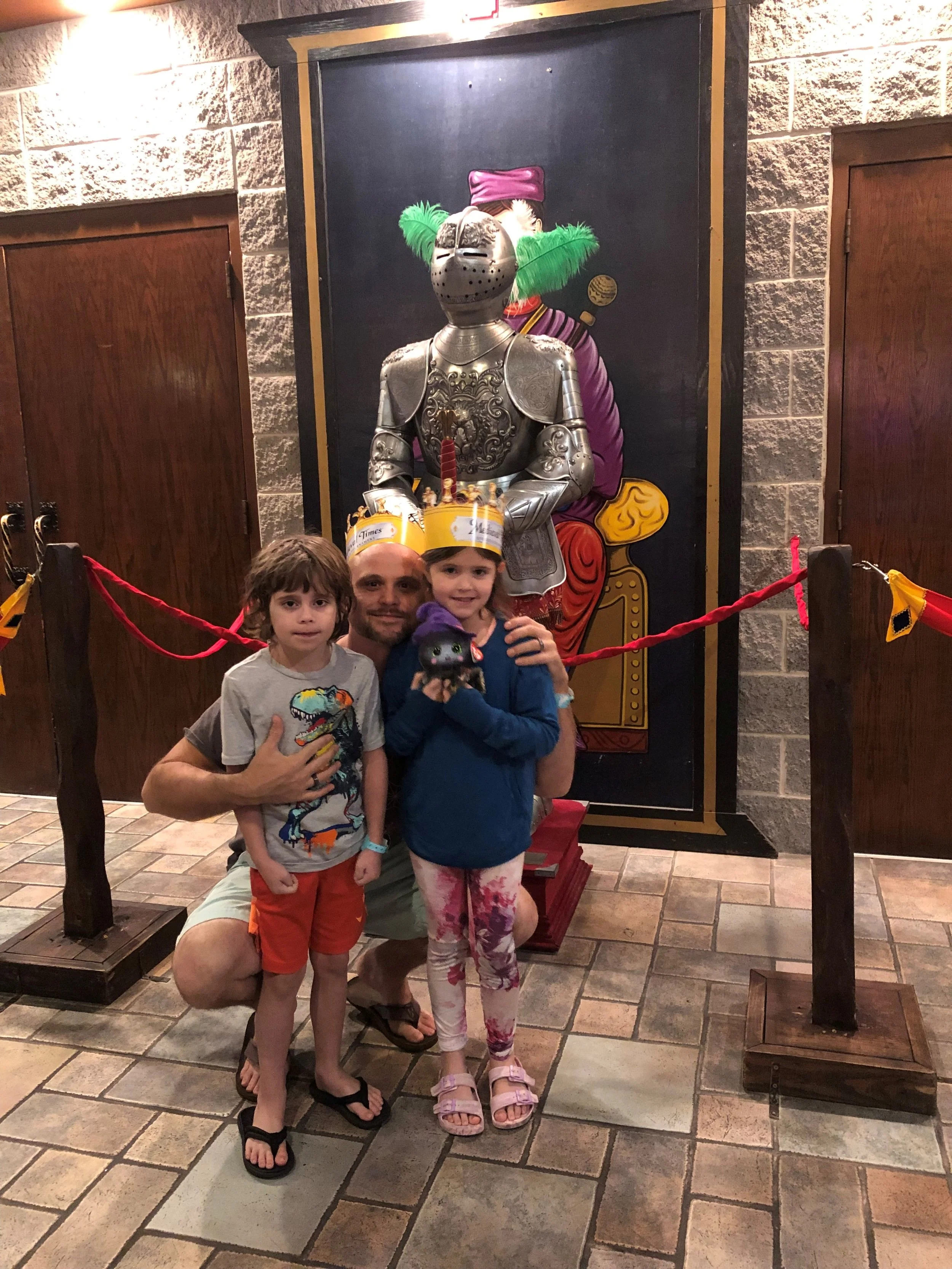 A man and two children posing in front of a knight statue at a themed attraction. The children are wearing matching 'Times' crowns, and the girl is holding a plush toy. An illustration of a knight on a throne is behind the statue.