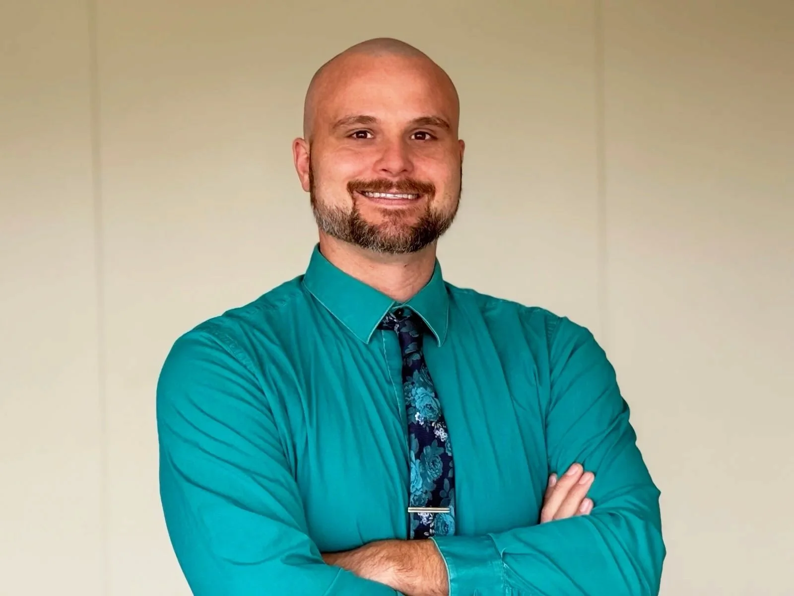 Portrait of a bald man with a beard, wearing a teal dress shirt and a patterned tie, standing with arms crossed and smiling against a neutral background.