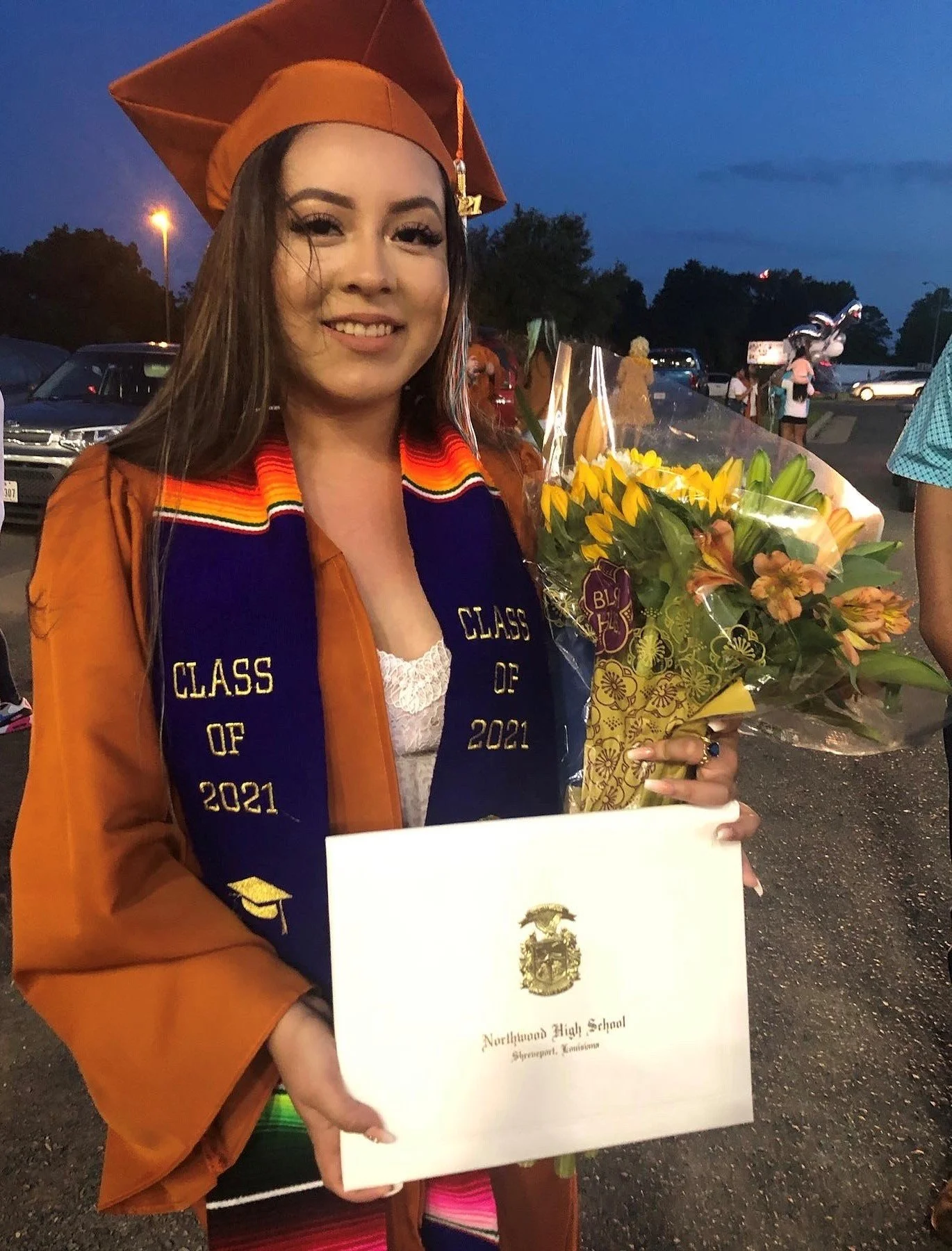 A young woman in a graduation cap and gown holding a bouquet of flowers and a diploma from Northland High School in Shreveport, Louisiana, celebrating graduation in the evening.