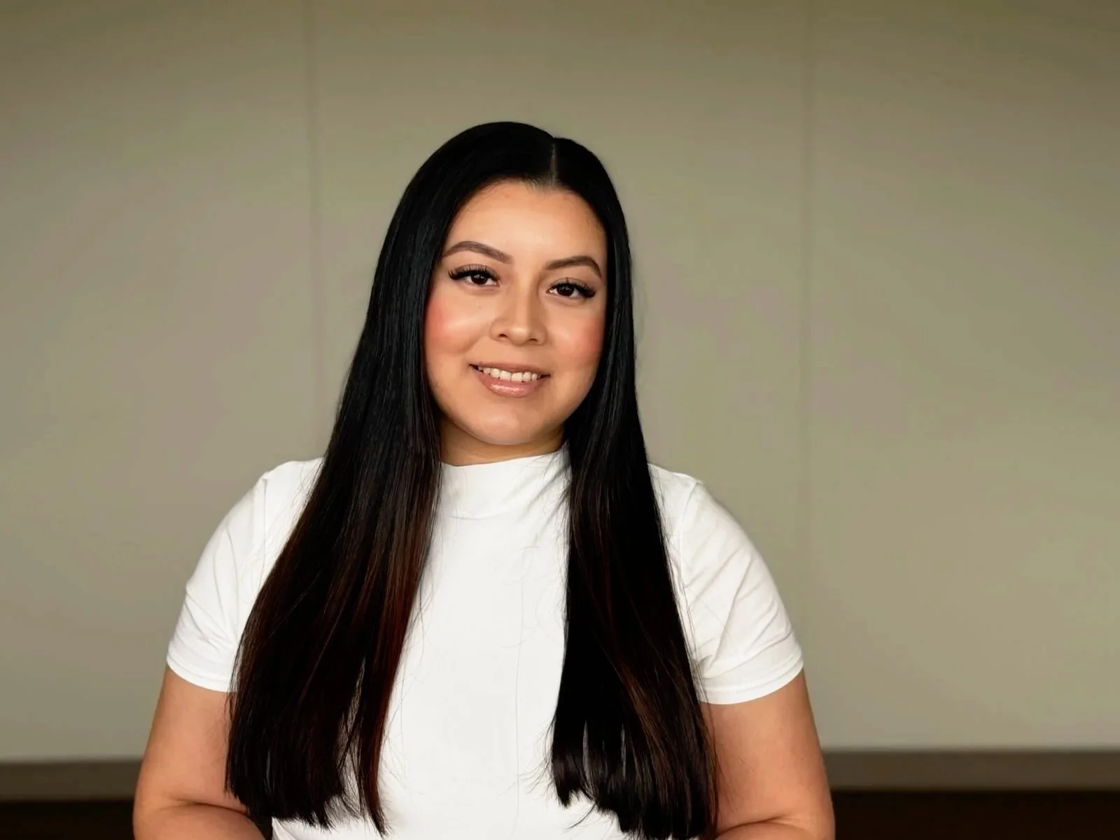 A woman with long black hair, wearing a white shirt, smiling at the camera in an indoor setting with a plain background.