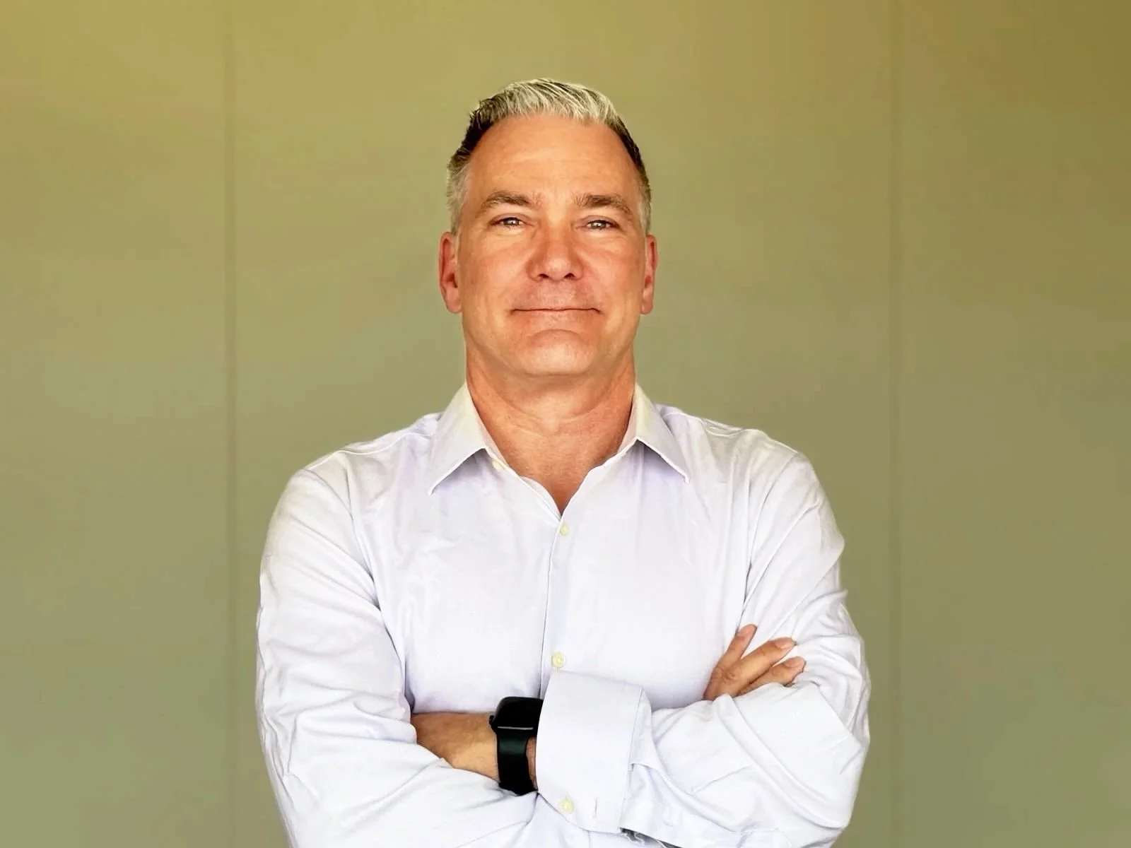 A middle-aged man with gray hair, wearing a white dress shirt and a black wristwatch, standing against a plain green wall with arms crossed, smiling slightly.