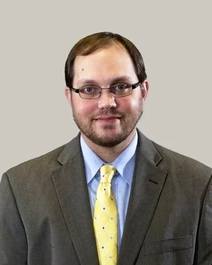 A man wearing glasses, a gray suit, a blue shirt, and a yellow tie, standing against a plain light gray background.