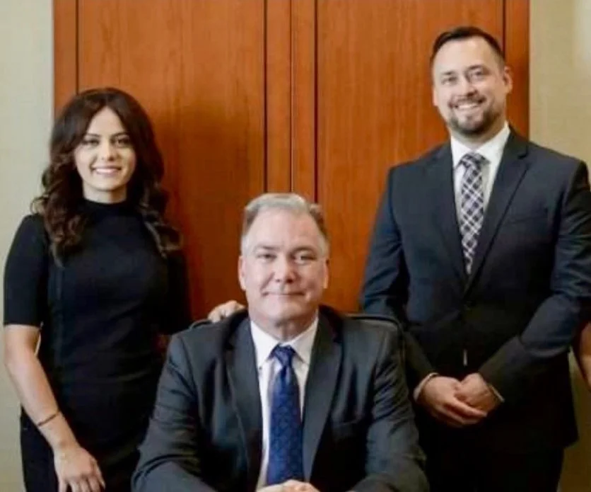 Three professionally dressed people posing for a photo in an office setting, with a man seated at the desk in the center and two standing behind him.