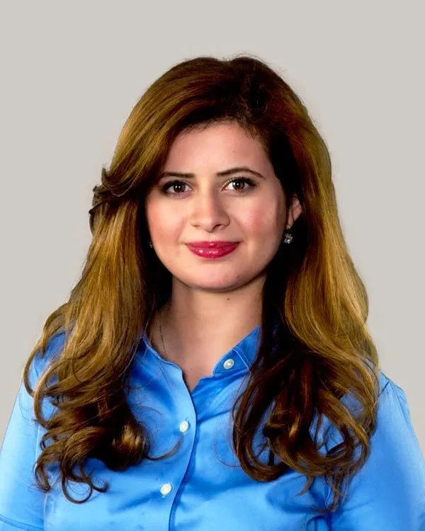A woman with long, wavy, light brown hair wearing a blue button-up shirt, smiling at the camera against a plain gray background.