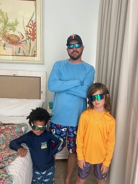 A man and two children wearing colorful sunglasses, standing in a bedroom with a bed, curtains, and artwork on the wall.